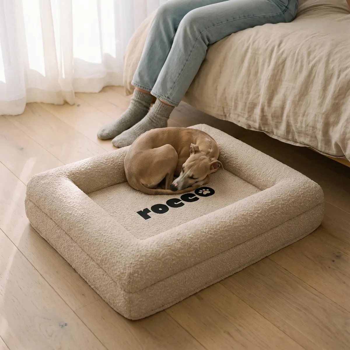 A whippet curled up asleep on an oatmeal Bolster Bed beside a bed